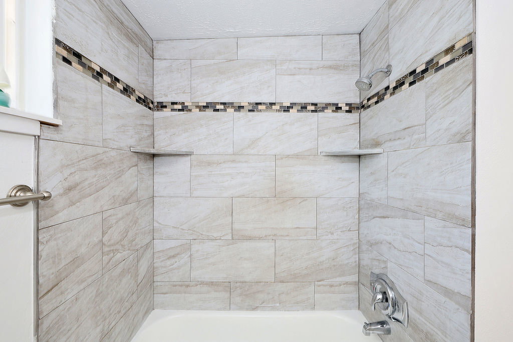 Bathtub shower with light-toned rectangular tile surround, horizontal mosaic accent band, and two built-in corner shelves