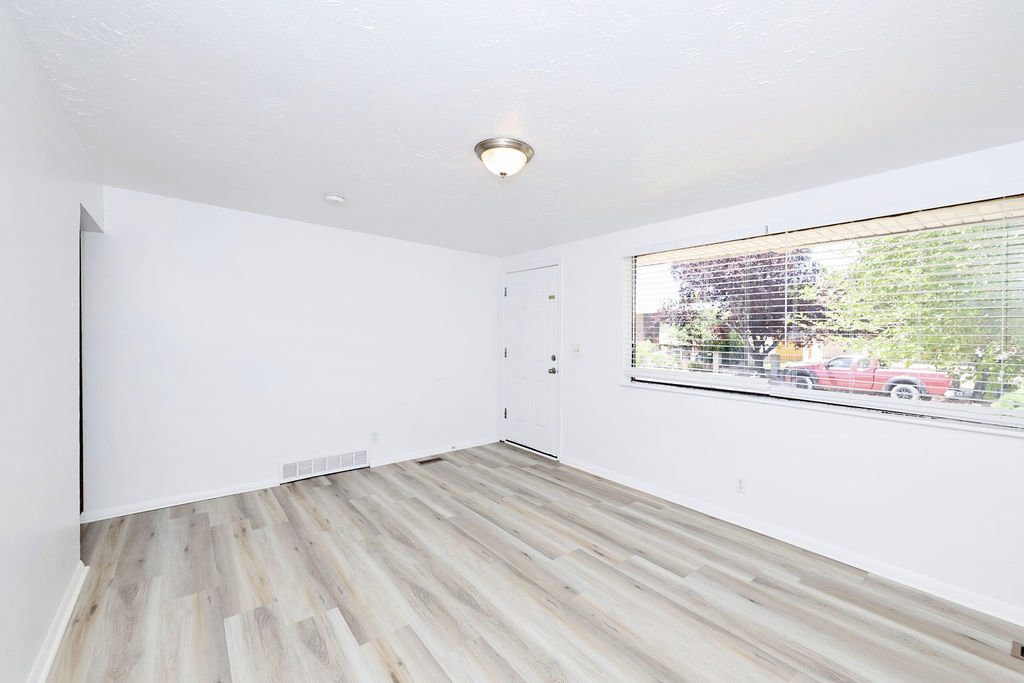 Spacious room with wood-finish flooring, white walls, a large window with blinds, and a ceiling-mounted light fixture