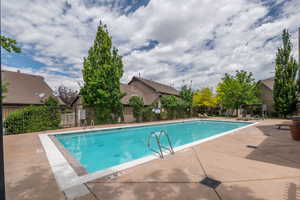 Sparkling community pool at ground level, surrounded by mature trees, iron fencing, lounge chairs, and a pergola seating area. Blue skies and clean water make this the place to be all summer.