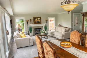 Spacious living area featuring a fireplace with a stone surround and white mantle, light gray wall paint, crown molding, and light-colored carpeting