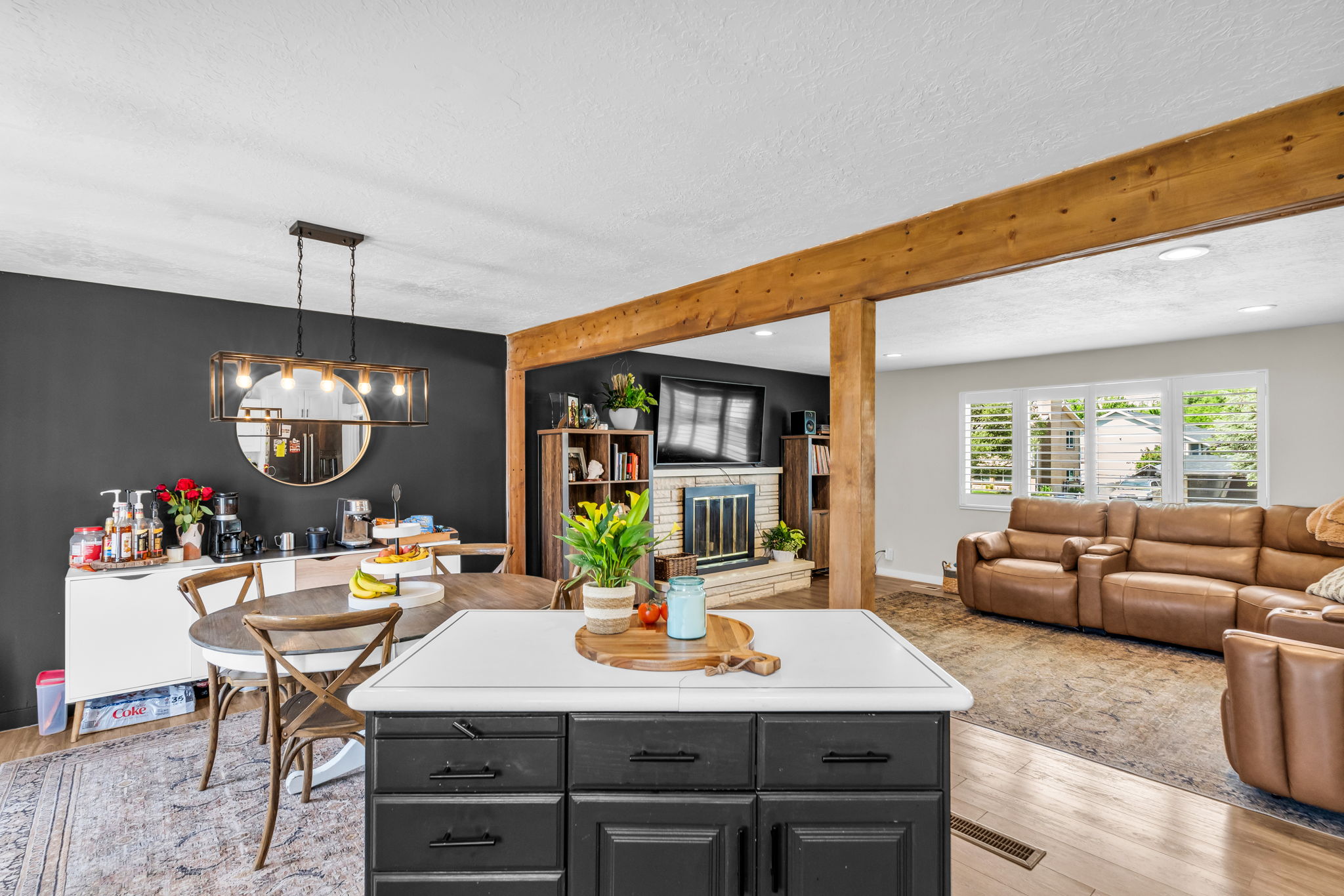 Open-concept living space with exposed wood beams, a central kitchen island with a white countertop, and a dining area featuring a modern linear chandelier