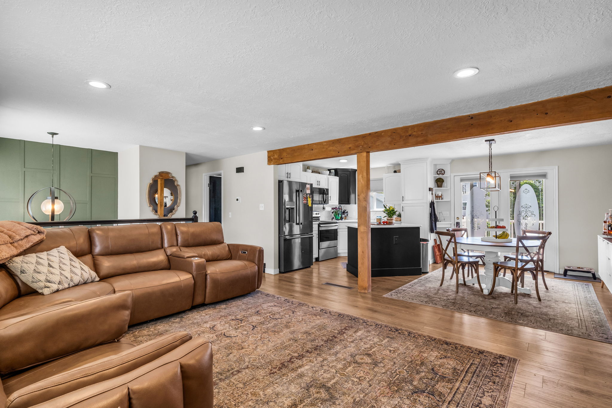 Open-concept living space featuring wood-finish flooring, a prominent exposed wood beam, recessed lighting, and a kitchen with dark appliances and white cabinetry