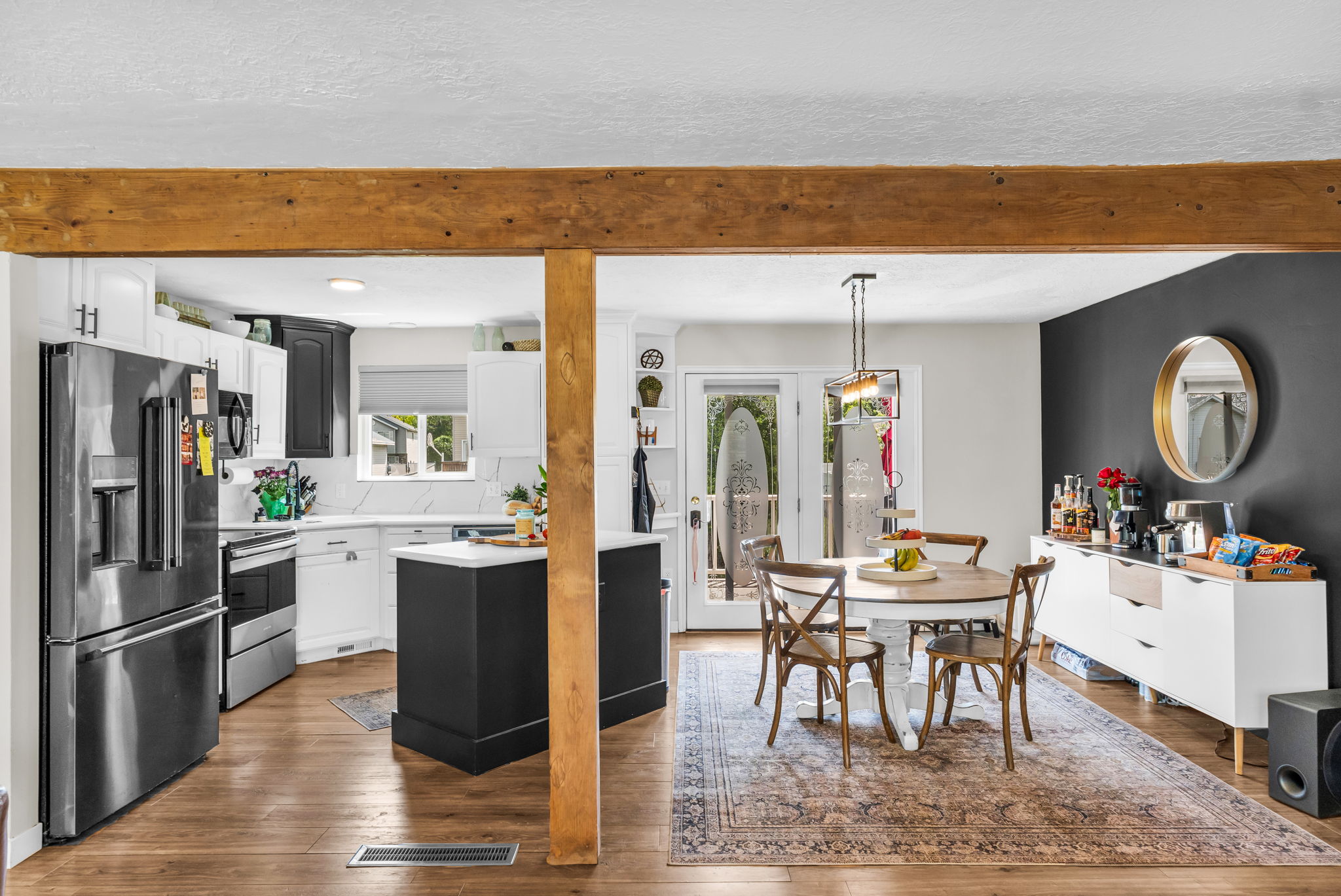 Open-concept kitchen and dining area featuring wood-finish flooring, exposed wood beam accents, a two-tone kitchen with white perimeter cabinetry and a dark island, stainless steel appliances, and double doors to an exterior space