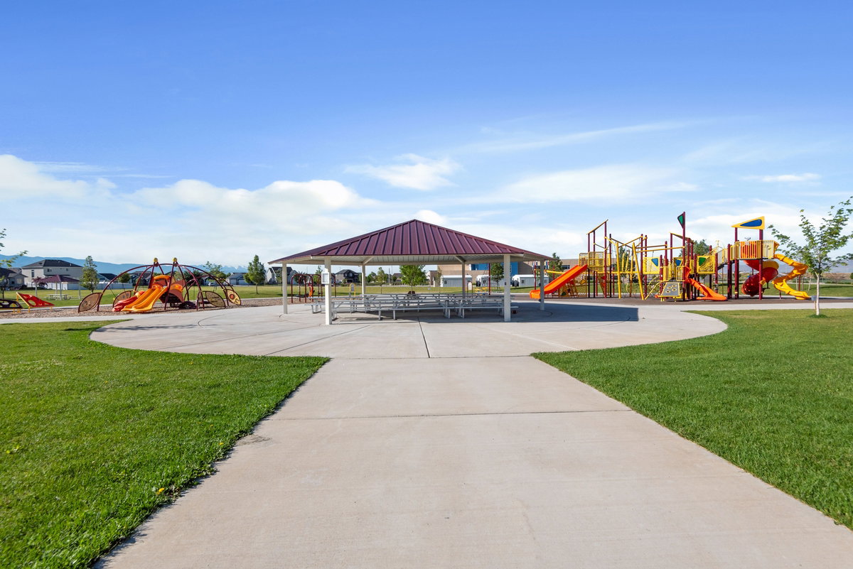 Expansive park grounds featuring a central covered pavilion, multiple playground structures, concrete pathways, and green lawn areas