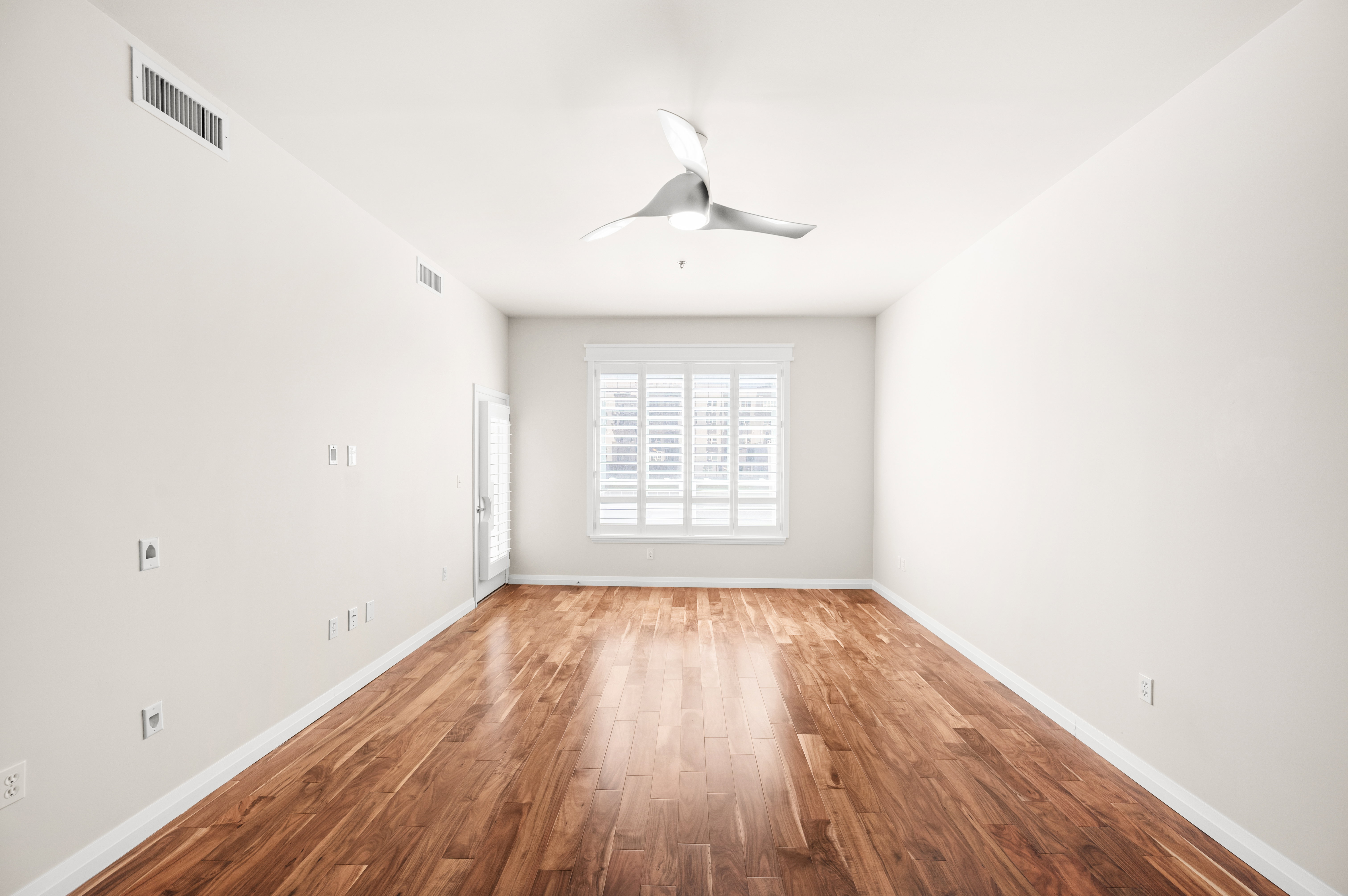 Spacious room featuring wood-finish flooring, a contemporary ceiling fan, and a window with plantation shutters