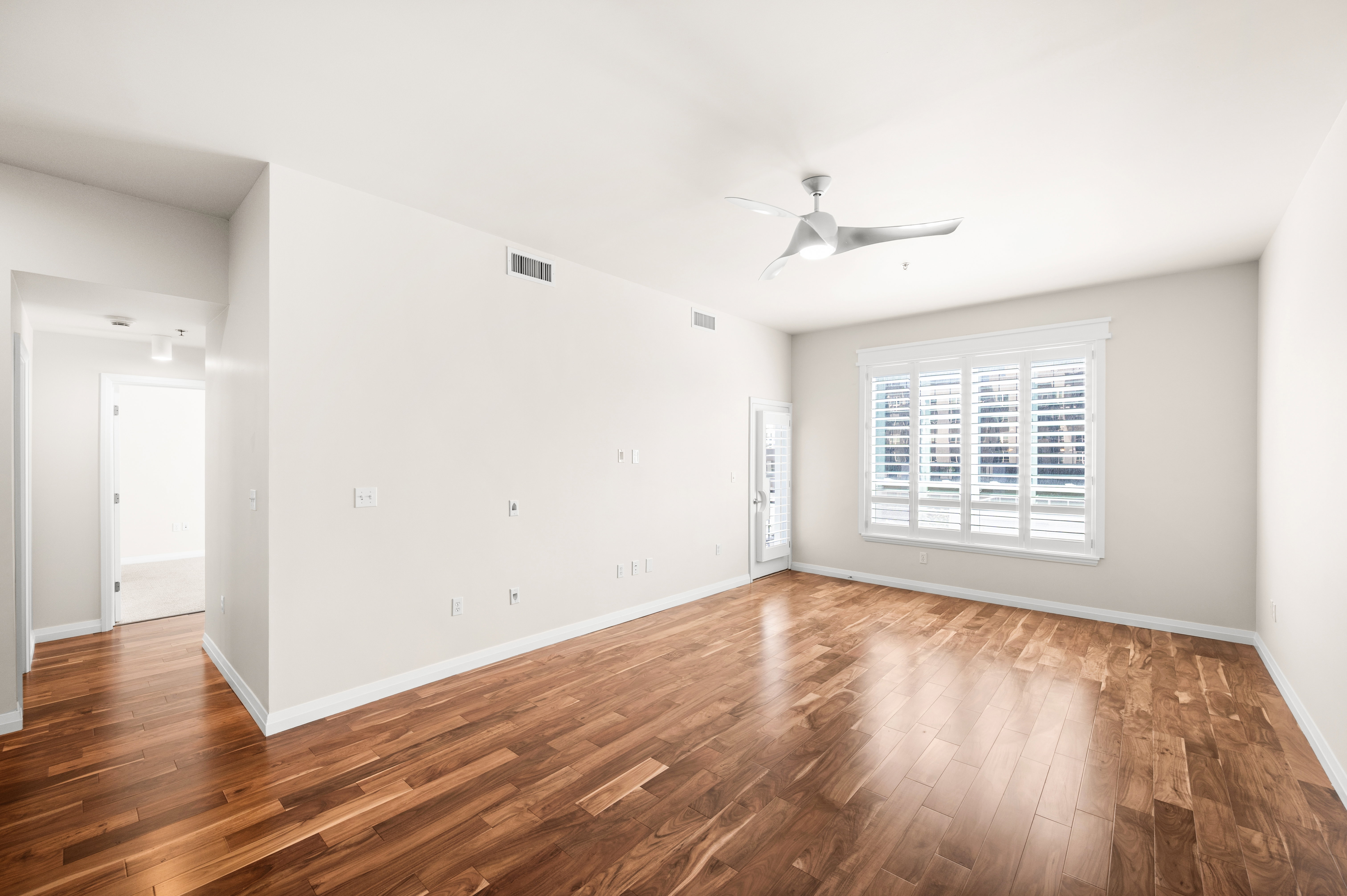 Spacious room featuring rich wood-finish flooring, a large window with white plantation shutters, a modern ceiling fan, recessed lighting, and a white interior door