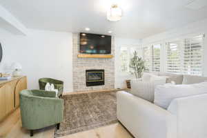 Living area featuring a stone fireplace with a wood-finish mantel, light wood-finish flooring, and white plantation shutters