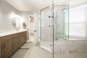 Spacious bathroom featuring a glass-enclosed shower with a hex-tiled floor, a built-in tub with a window, a double vanity with a light wood-finish cabinet, and light-toned tile flooring