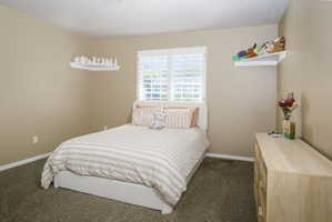 Bedroom featuring tan wall paint, textured carpet flooring, white window shutters, and white baseboards