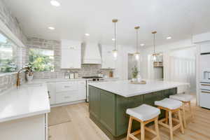 Modern kitchen featuring a large central island with a white countertop and olive green base, light wood-finish flooring, white cabinetry, integrated stainless steel appliances, and a full-height stone backsplash