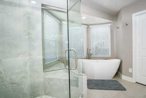 Contemporary bathroom featuring a freestanding soaking tub, separate glass-enclosed shower with tile surround, bay windows with blinds, and light-toned tile flooring