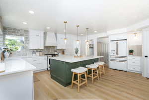 Spacious kitchen featuring a large green island with seating, white shaker cabinetry, stone backsplash, wood-finish flooring, and integrated appliances