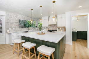 Kitchen featuring a large island with a white countertop and green paneling, light wood-finish flooring, white upper and lower cabinetry, stone-finish backsplash, and three brass-finish pendant lights