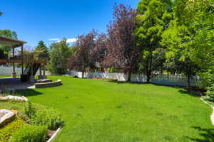 Expansive outdoor living area featuring a large, level lawn, a multi-level wood-finish deck with black metal railings, a stone-finish patio, a built-in stone-finish seating wall, and a white picket fence