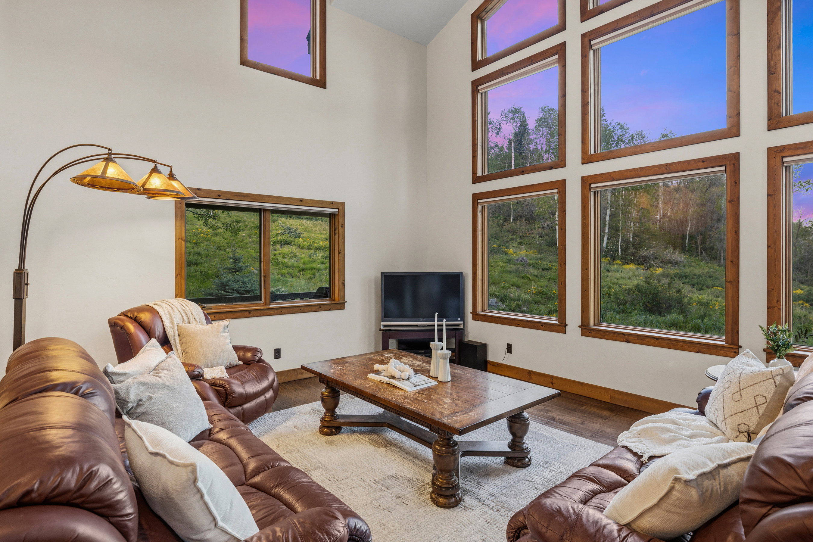 Two-story vaulted ceiling living area featuring a wall of multiple large windows, wood-finish flooring, and a substantial wood-frame window system