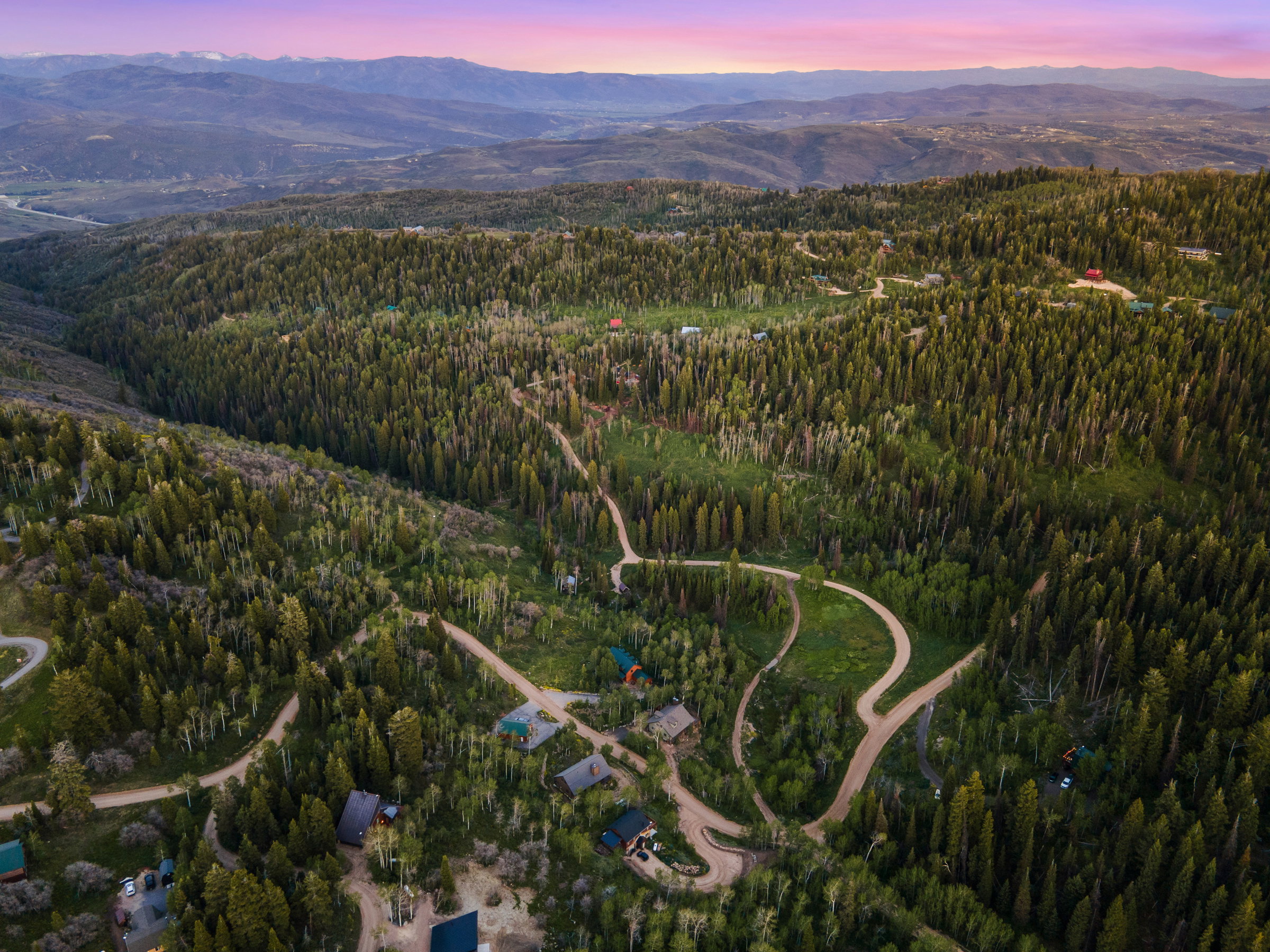 Expansive mountainous landscape featuring extensive evergreen forests, winding roads, and distant mountain ranges under a colorful sky
