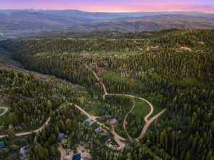 Expansive mountainous landscape featuring extensive evergreen forests, winding roads, and distant mountain ranges under a colorful sky