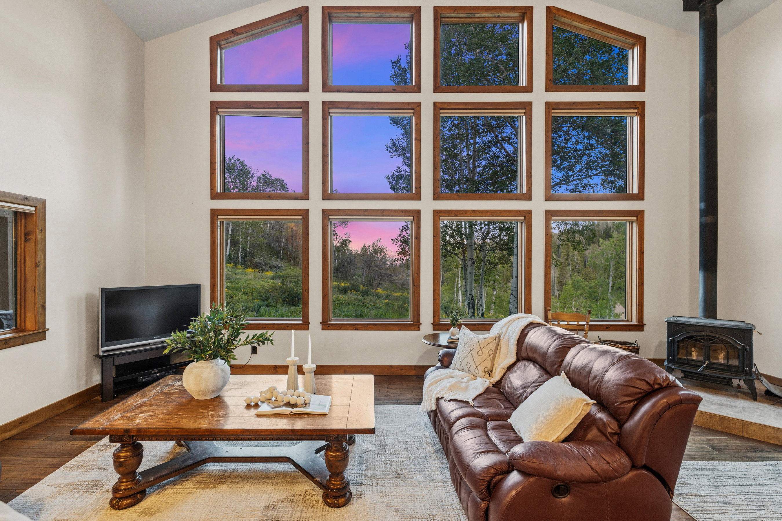 Vaulted ceiling living space featuring a wall of wood-framed windows, wood-finish flooring, and a black wood-burning stove with a stovepipe