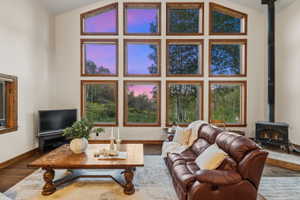 Vaulted ceiling living space featuring a wall of wood-framed windows, wood-finish flooring, and a black wood-burning stove with a stovepipe