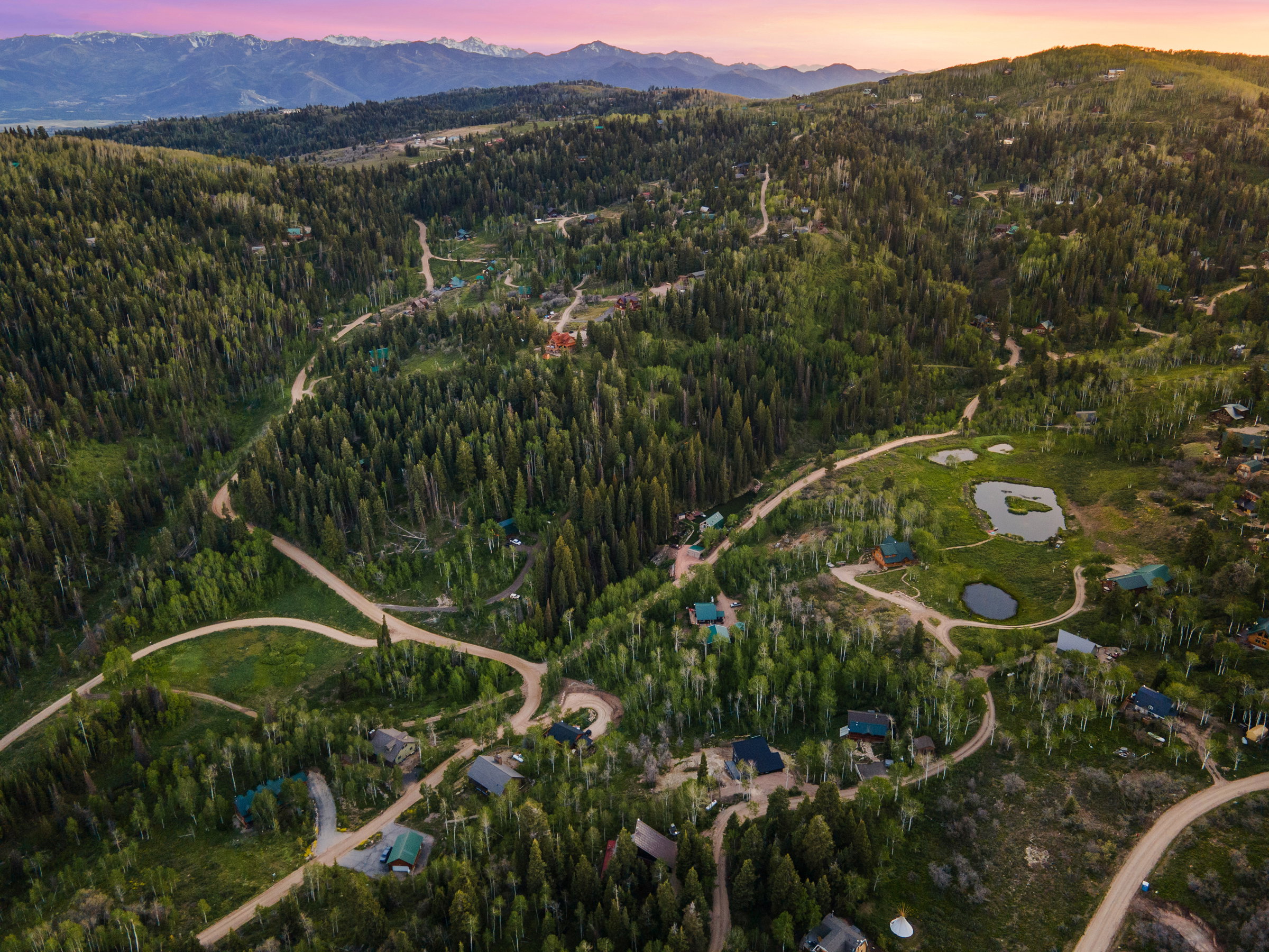 Expansive mountain terrain featuring dense evergreen and deciduous trees, multiple private residences, winding unpaved roads, and several natural ponds