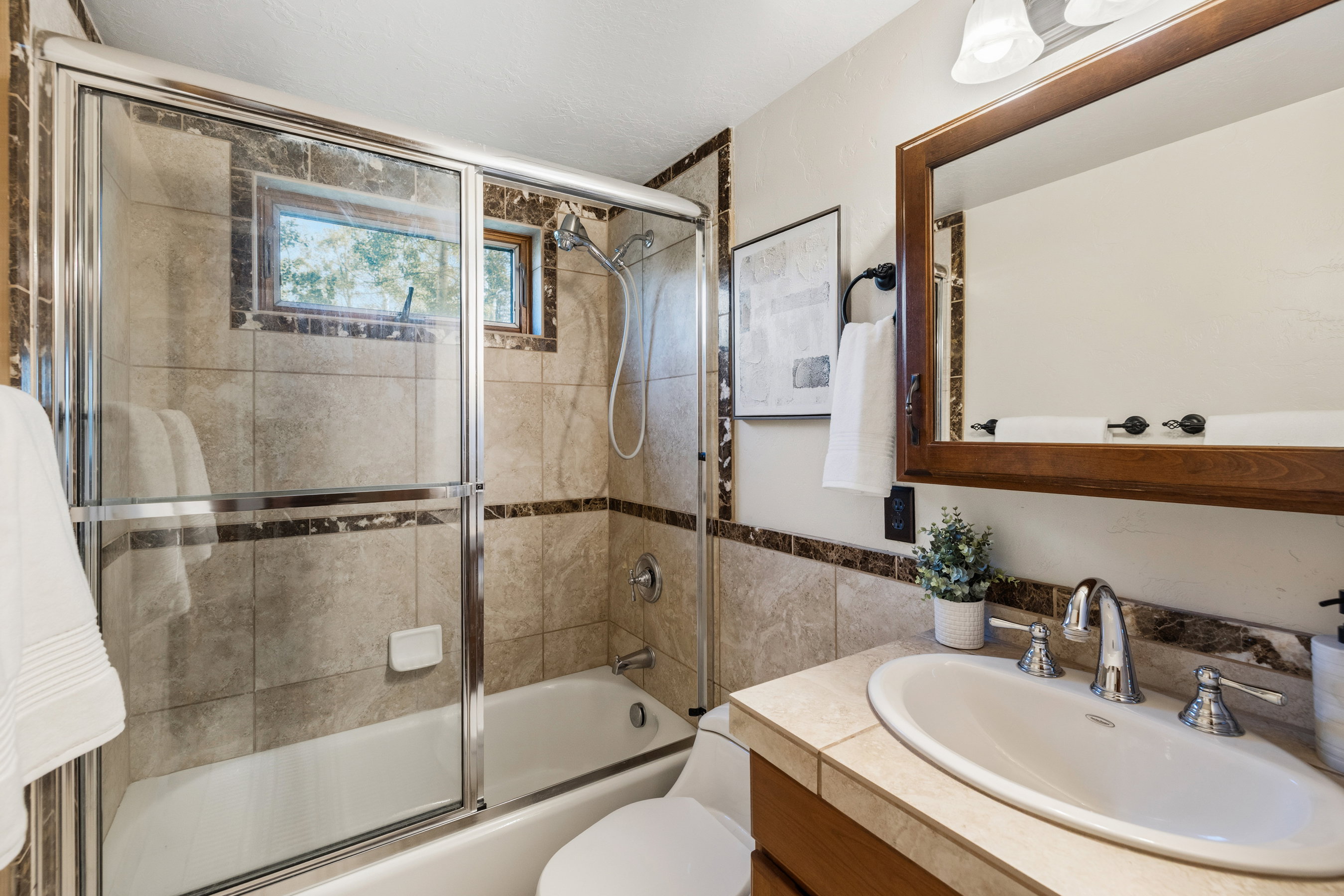 Bathroom featuring a tiled shower/tub combination with glass enclosure, a window, a wood-framed mirror, and a vanity with an integrated sink and chrome fixtures