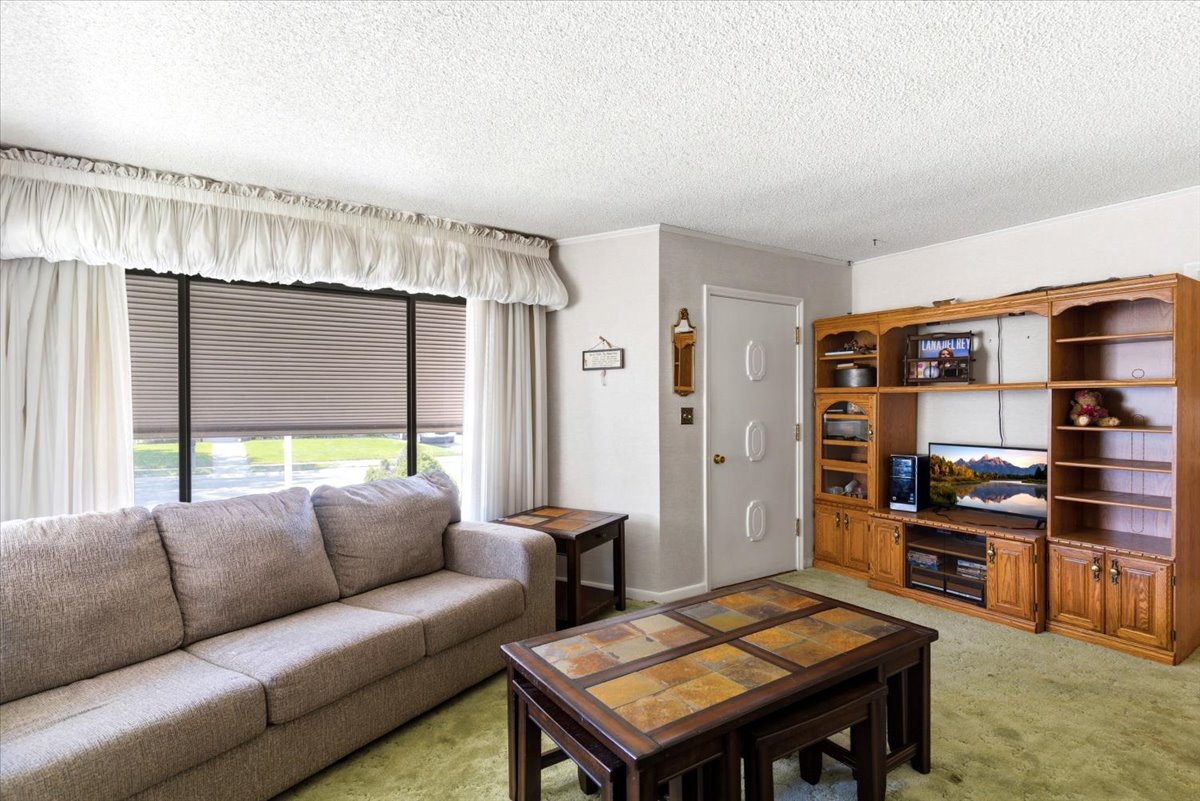 Spacious living area featuring a large window with horizontal blinds, light-colored walls, and green carpet flooring