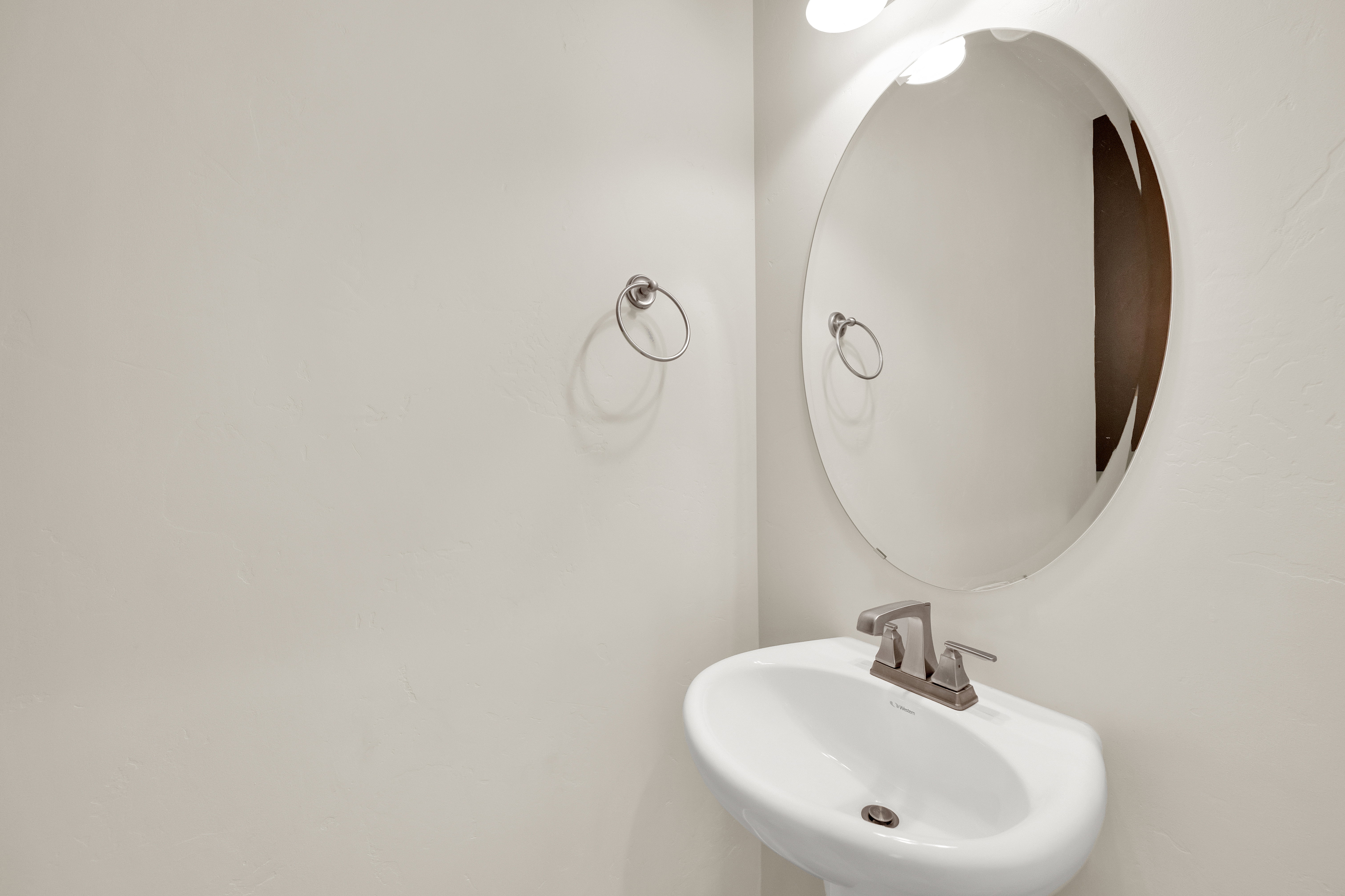 Pedestal sink with a brushed nickel faucet, an oval frameless mirror, and a wall-mounted light fixture
