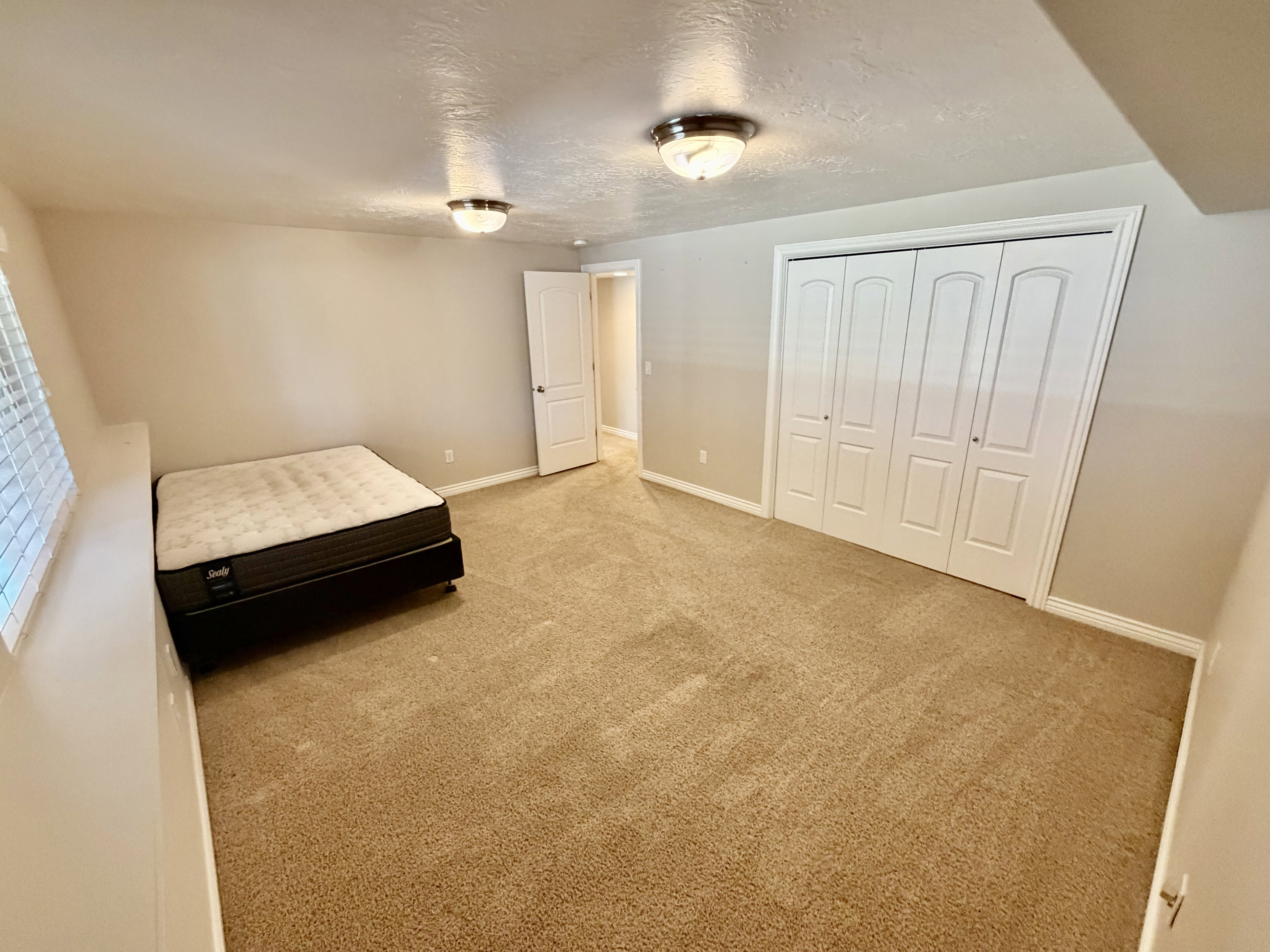 Spacious room featuring neutral carpeting, a window with horizontal blinds, and a built-in closet with bifold doors