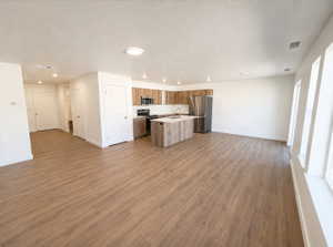Open-concept living space featuring wood-finish flooring, a kitchen island with sink, stainless steel appliances, and multiple windows