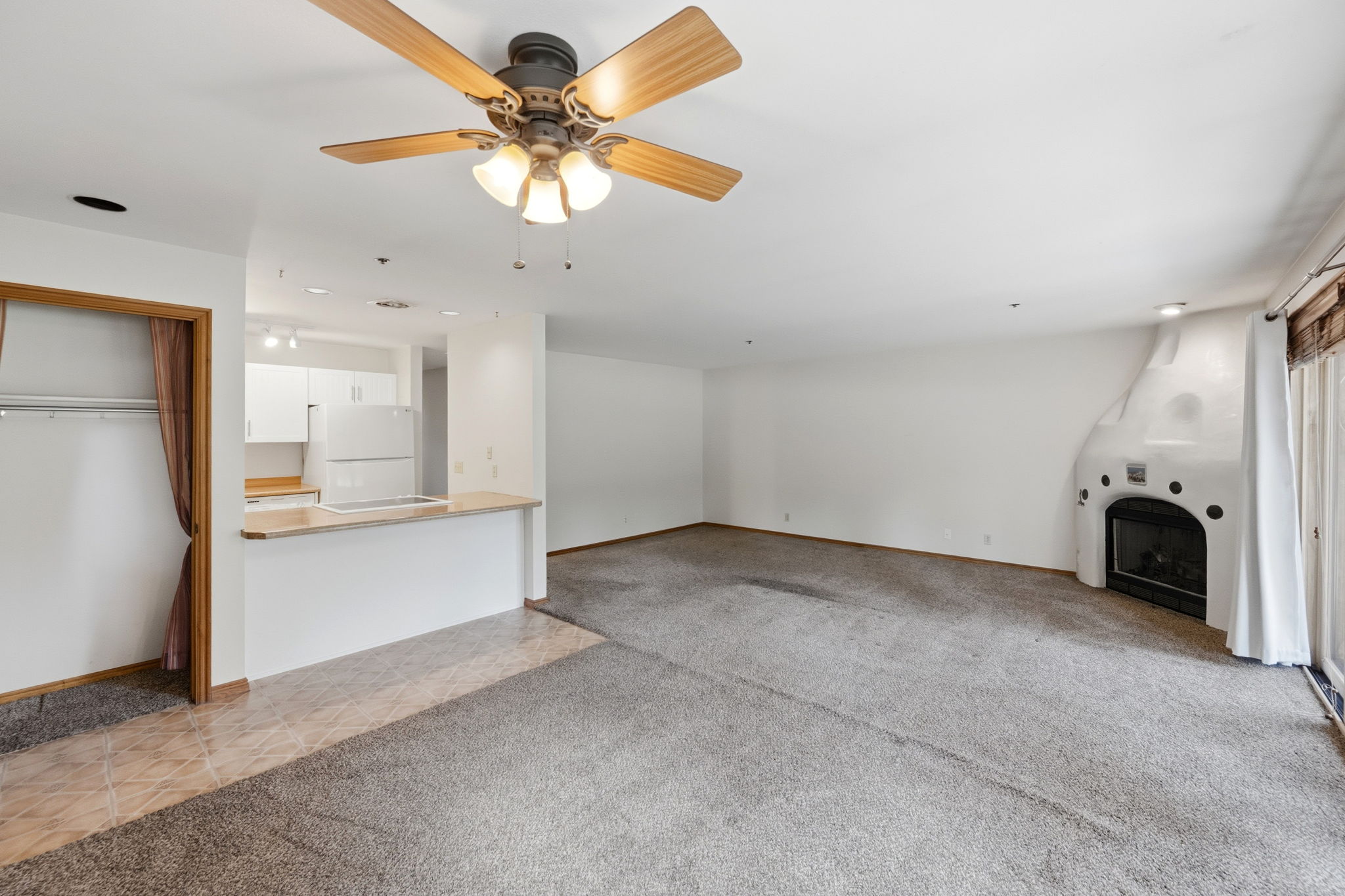 Living area featuring a textured carpet, a kiva-style fireplace, and a ceiling fan with integrated lighting