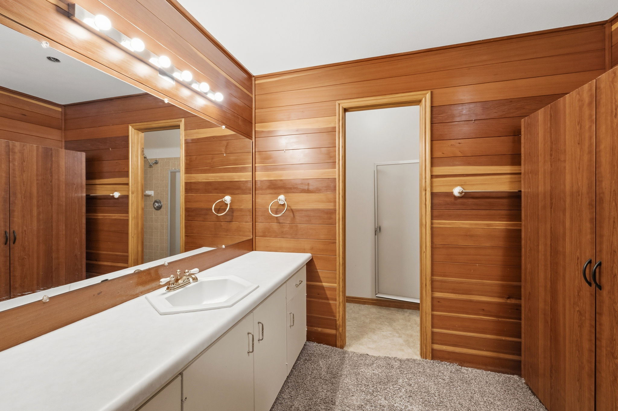 Bathroom featuring extensive wood paneling, a full vanity with an integrated sink, a large mirror with strip lighting, and a separate shower area