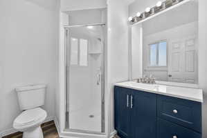 Bathroom featuring a dark blue vanity with a white countertop, chrome hardware, a glass-enclosed shower, a white toilet, and wood-finish flooring