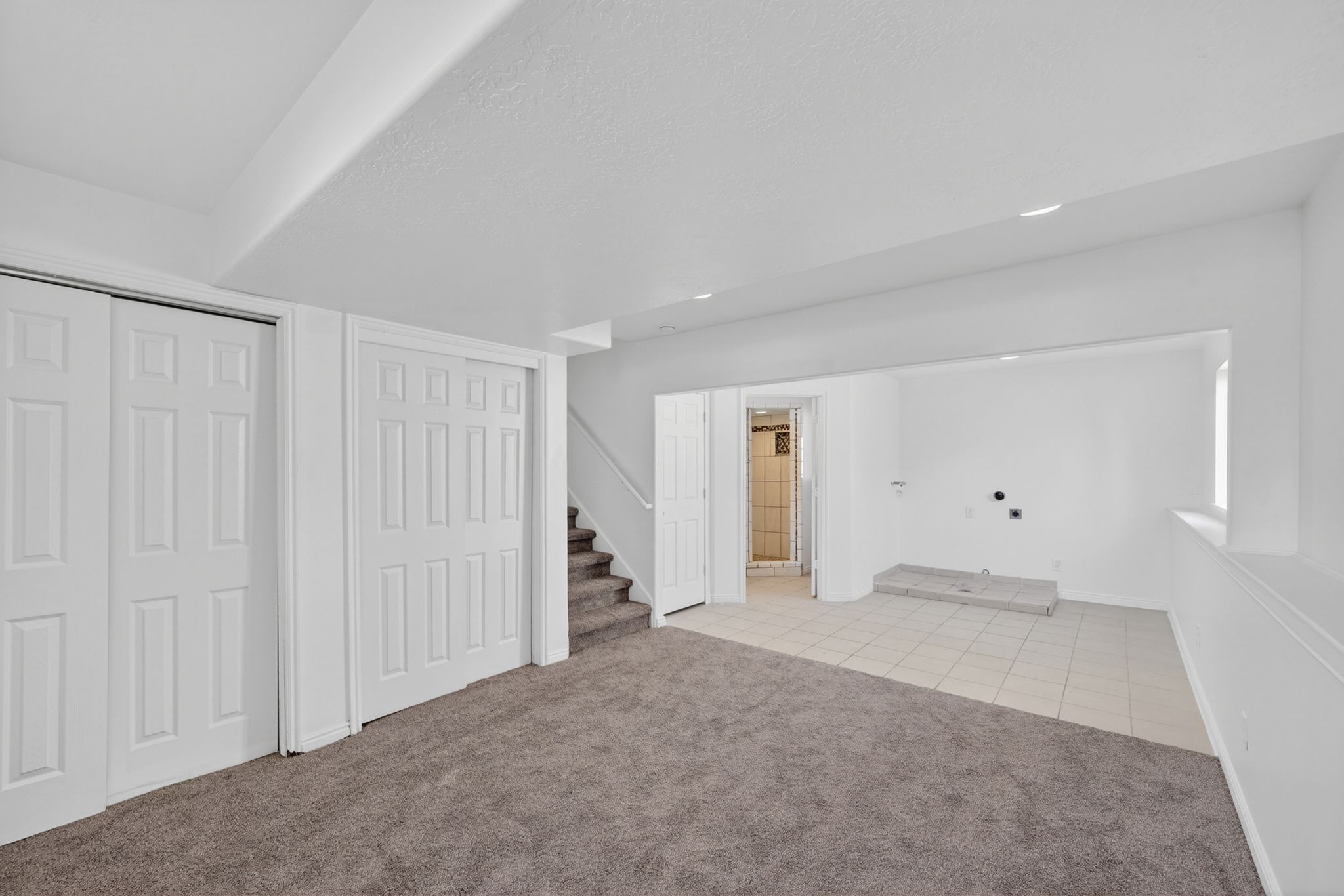Carpeted open-concept room featuring paneled bi-fold closet doors, a staircase with carpeted treads, and a tiled utility area with visible plumbing connections