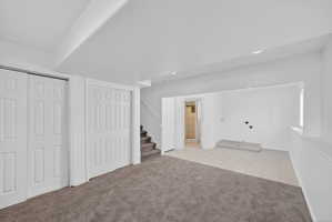 Carpeted open-concept room featuring paneled bi-fold closet doors, a staircase with carpeted treads, and a tiled utility area with visible plumbing connections