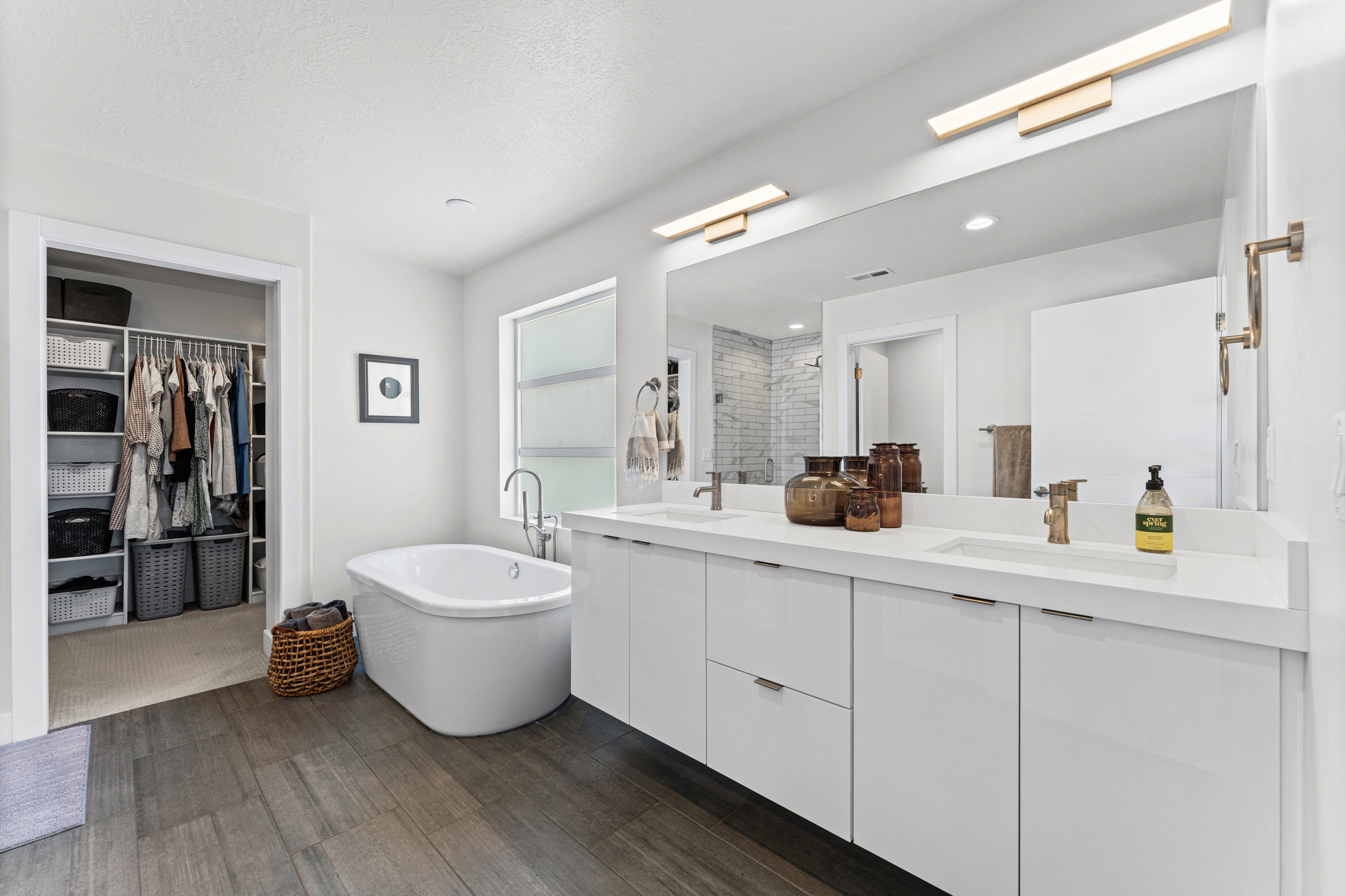 Contemporary bathroom featuring a freestanding soaking tub, wood-finish flooring, a double vanity with white cabinetry, and a large wall-mounted mirror with integrated lighting