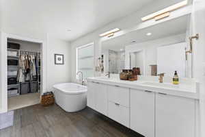Contemporary bathroom featuring a freestanding soaking tub, wood-finish flooring, a double vanity with white cabinetry, and a large wall-mounted mirror with integrated lighting