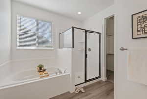 White soaking tub with brushed nickel fixtures, a window with blinds, a separate shower with a black-framed glass enclosure, and wood-finish flooring