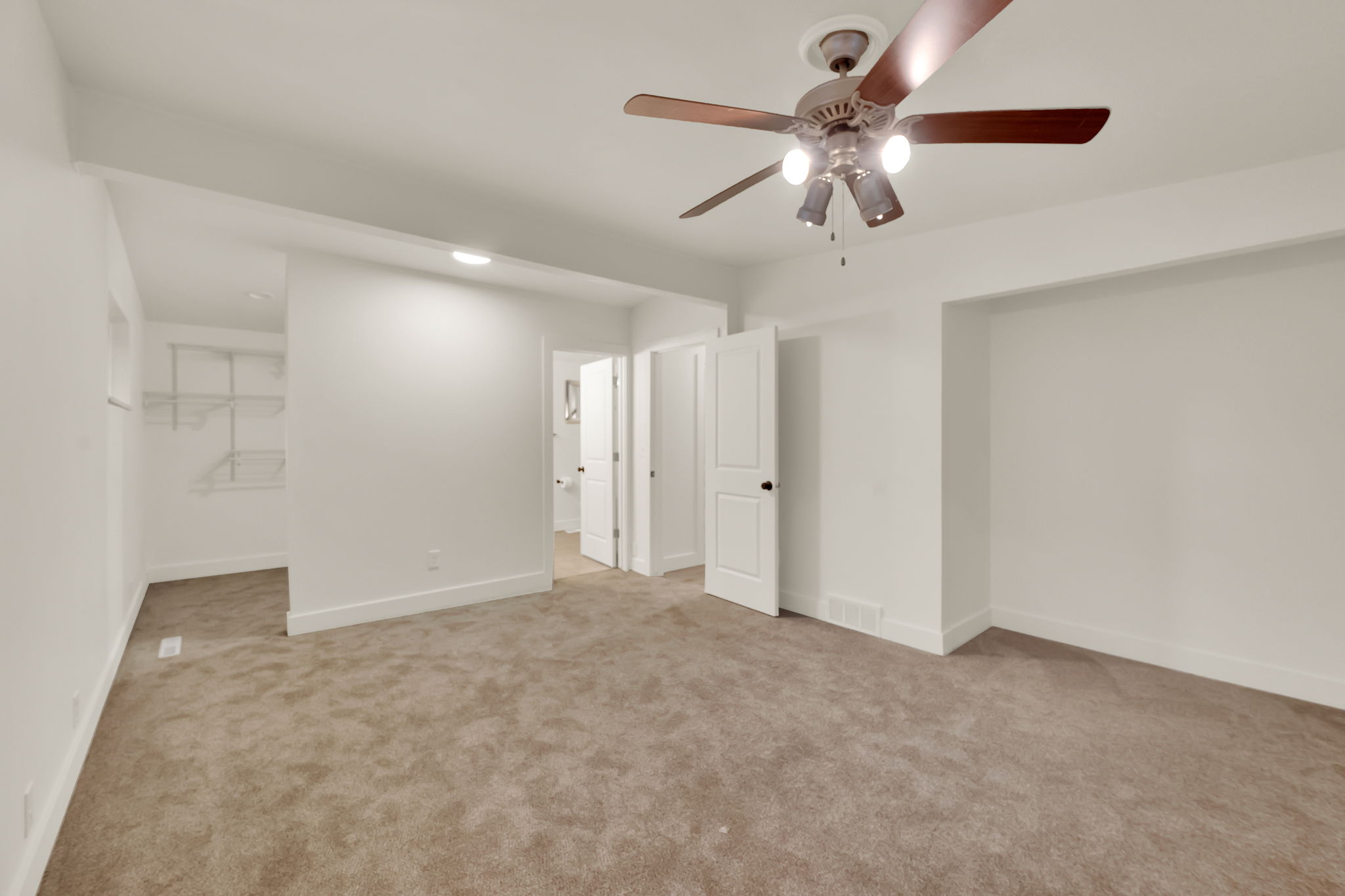 Spacious carpeted room featuring a ceiling fan with integrated lighting, a built-in open closet with wire shelving, and multiple white paneled doors