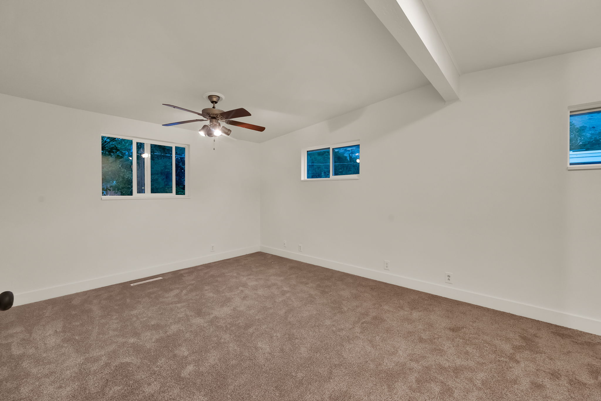 Spacious interior room featuring plush carpeting, multiple windows, a ceiling fan with integrated lighting, and a prominent ceiling beam