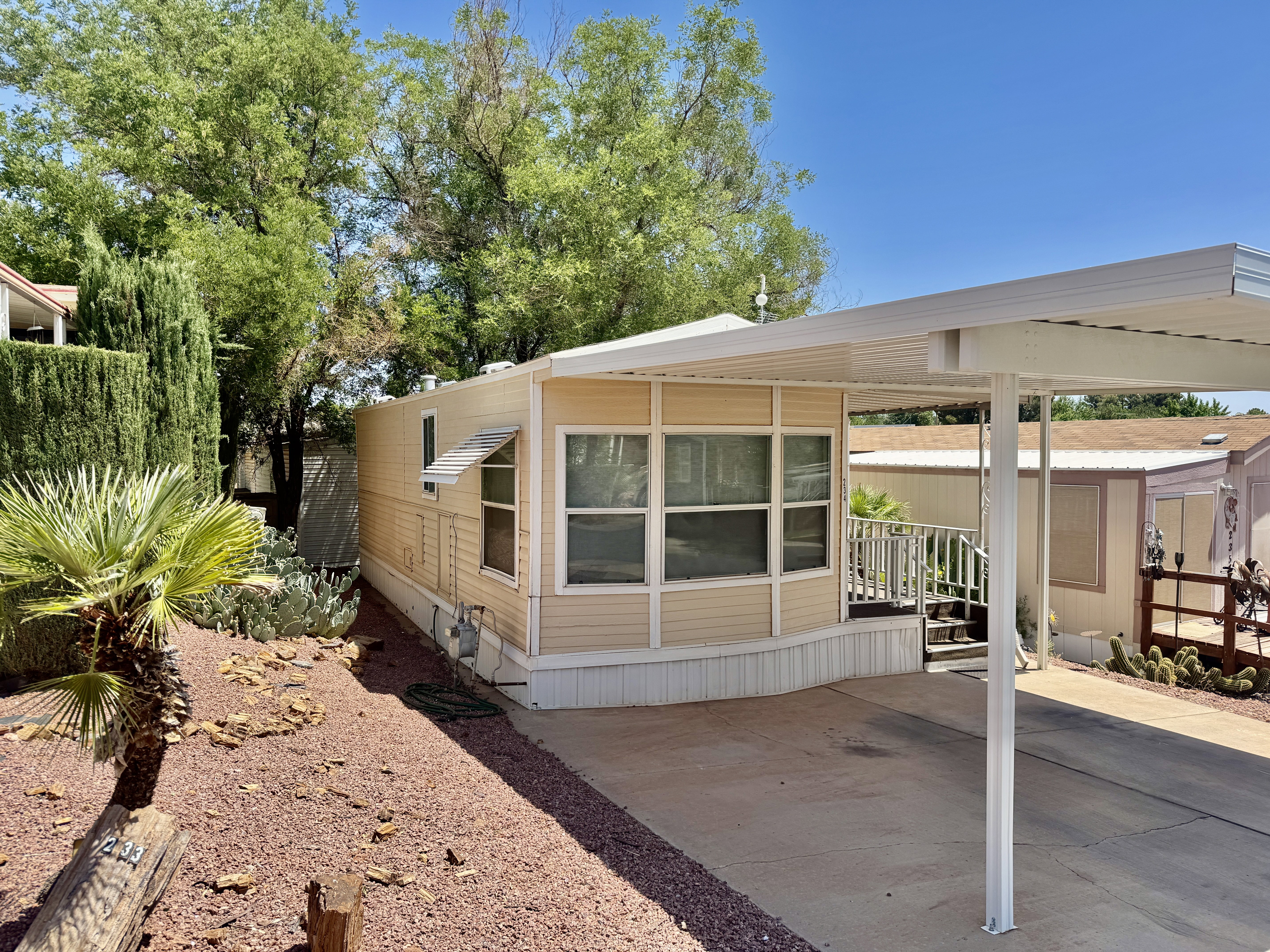 Beige exterior siding with a covered carport, rock landscaping, mature trees, and an elevated entry with white railings