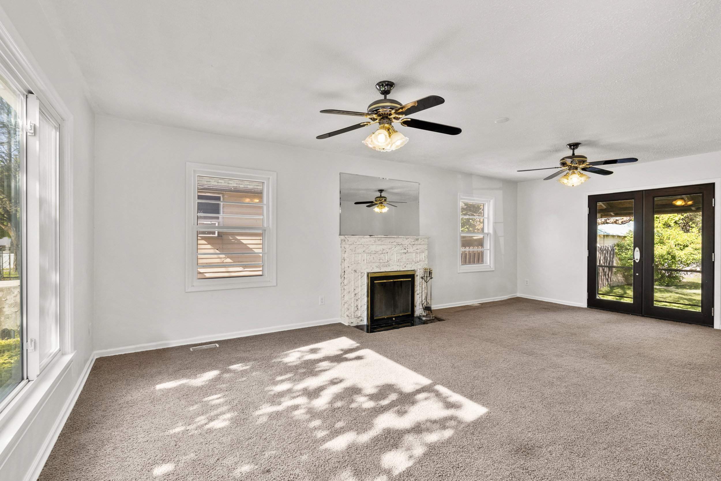Spacious interior room featuring light-toned carpeting, a white brick fireplace with a dark firebox, and two ceiling fans with integrated lighting