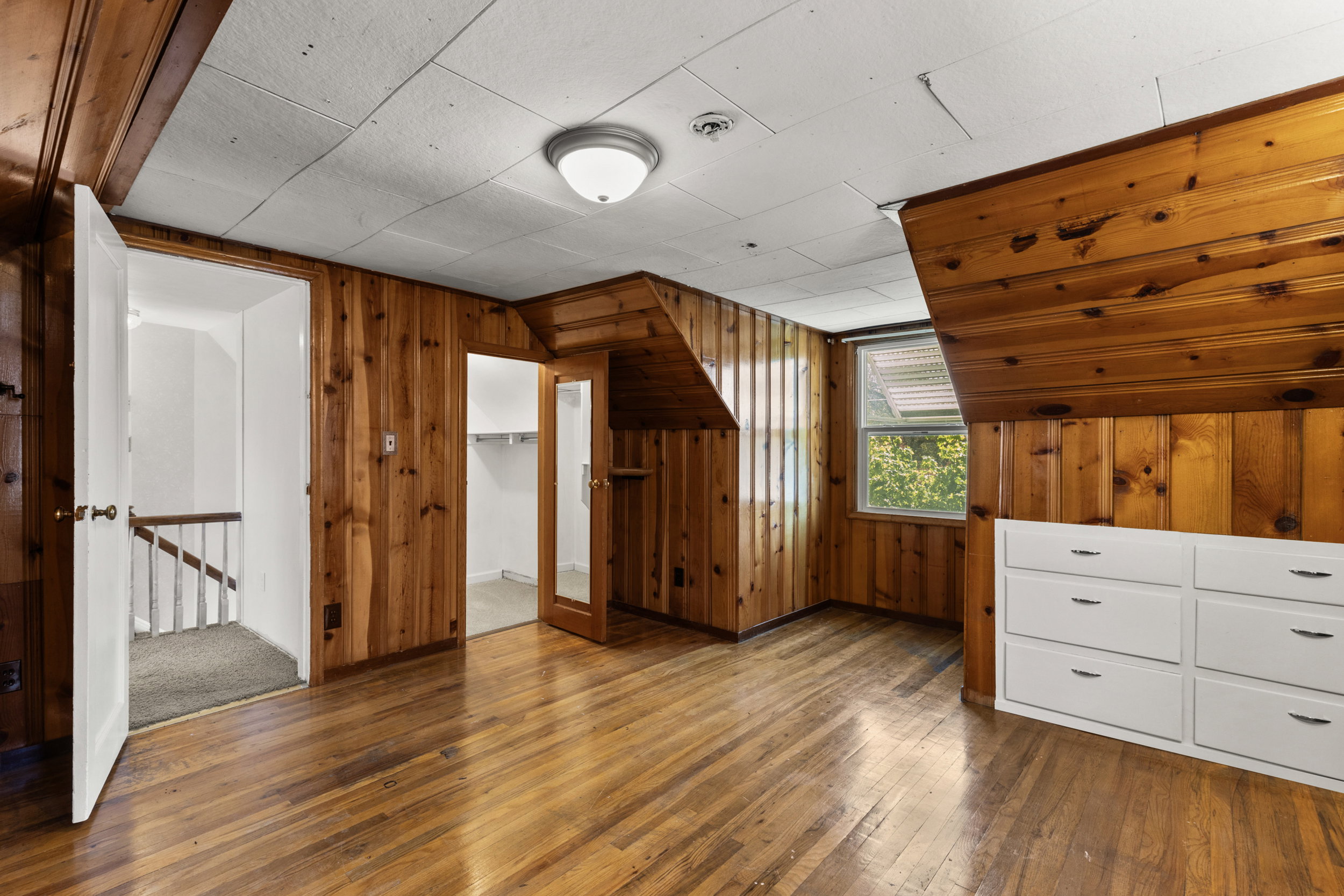 Wood-paneled room featuring wood-finish flooring, a window providing natural light, a built-in white dresser with six drawers, and a ceiling-mounted light fixture