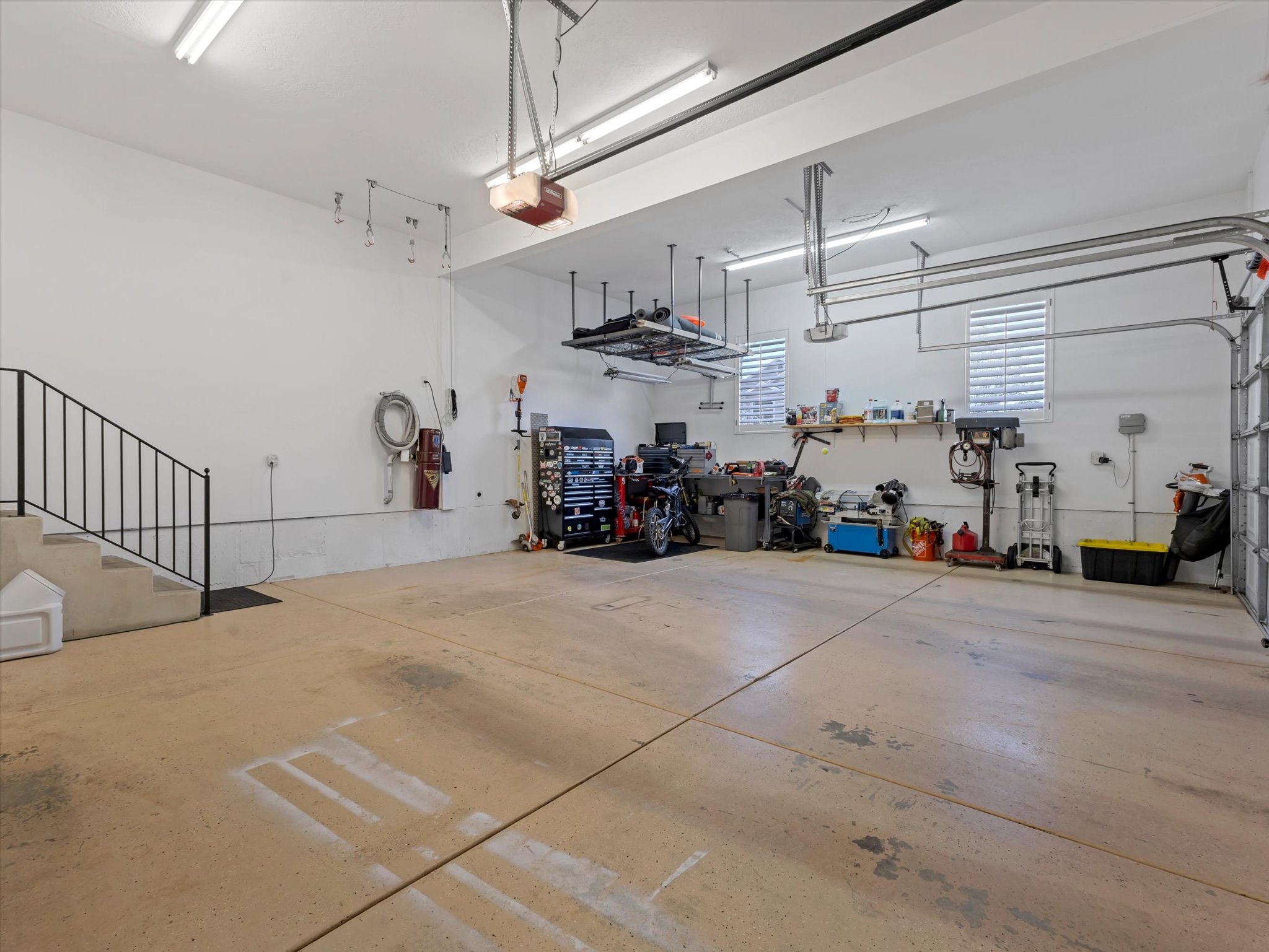 Spacious two-car garage featuring a concrete floor, built-in shelving, abundant overhead lighting, and two windows with horizontal blinds