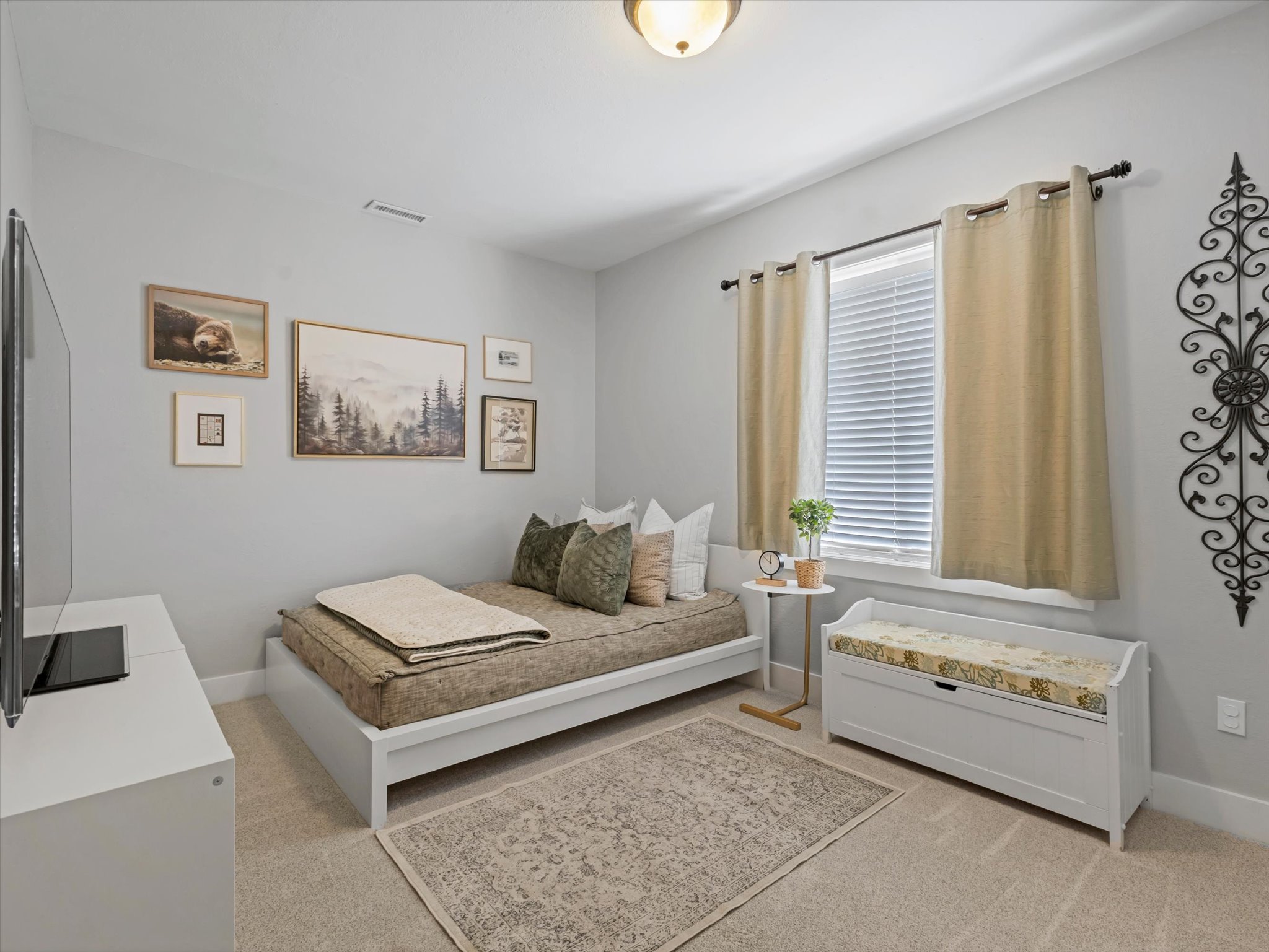 Carpeted room featuring a single window with blinds and drapes, a built-in ceiling light fixture, a wall-mounted decorative metal scroll, and a white storage bench with a patterned cushion