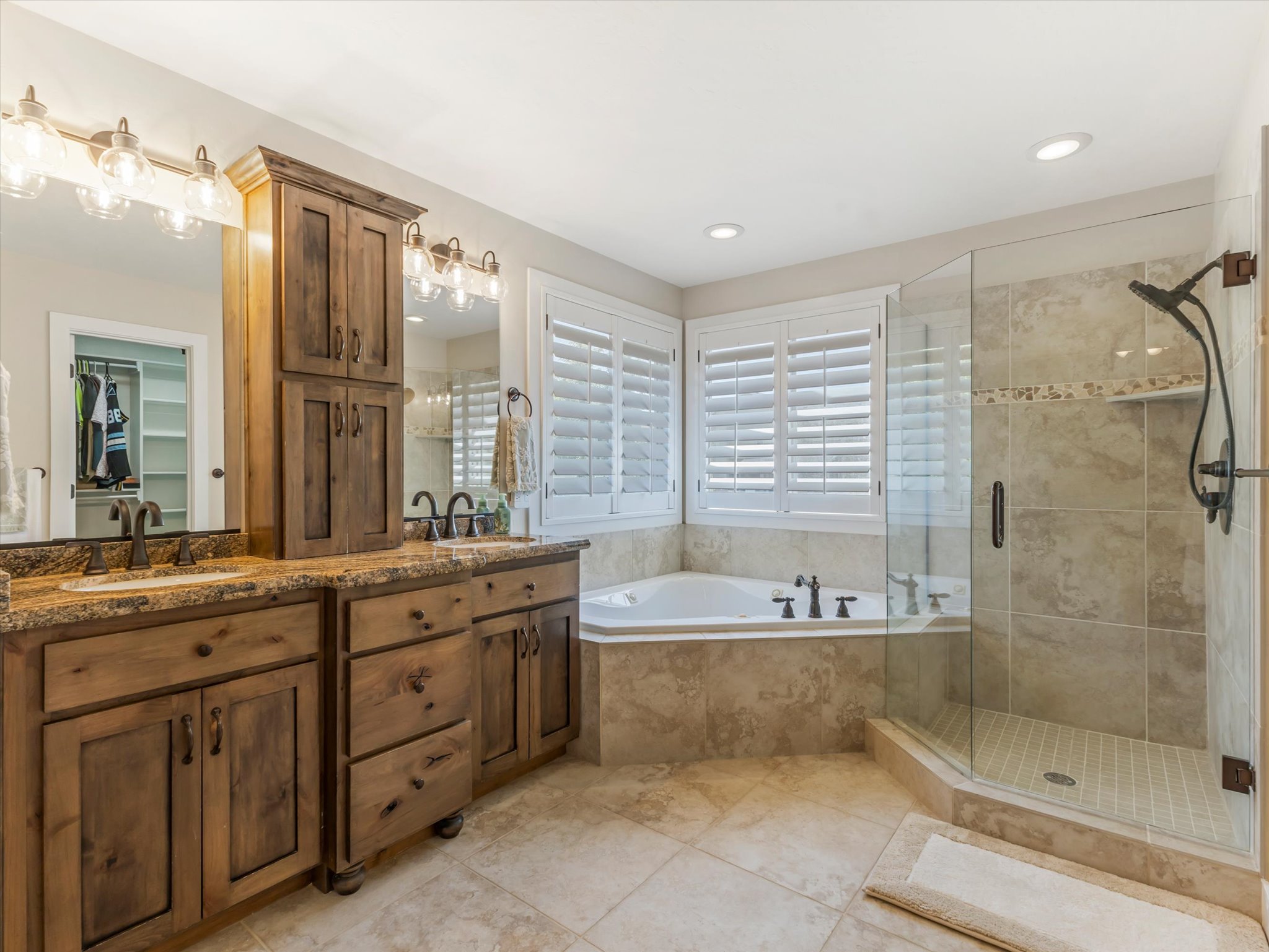 Bathroom featuring a dual vanity with wood cabinetry and stone countertops, a deep soaking tub, a glass-enclosed shower with tile surround, and plantation shutters