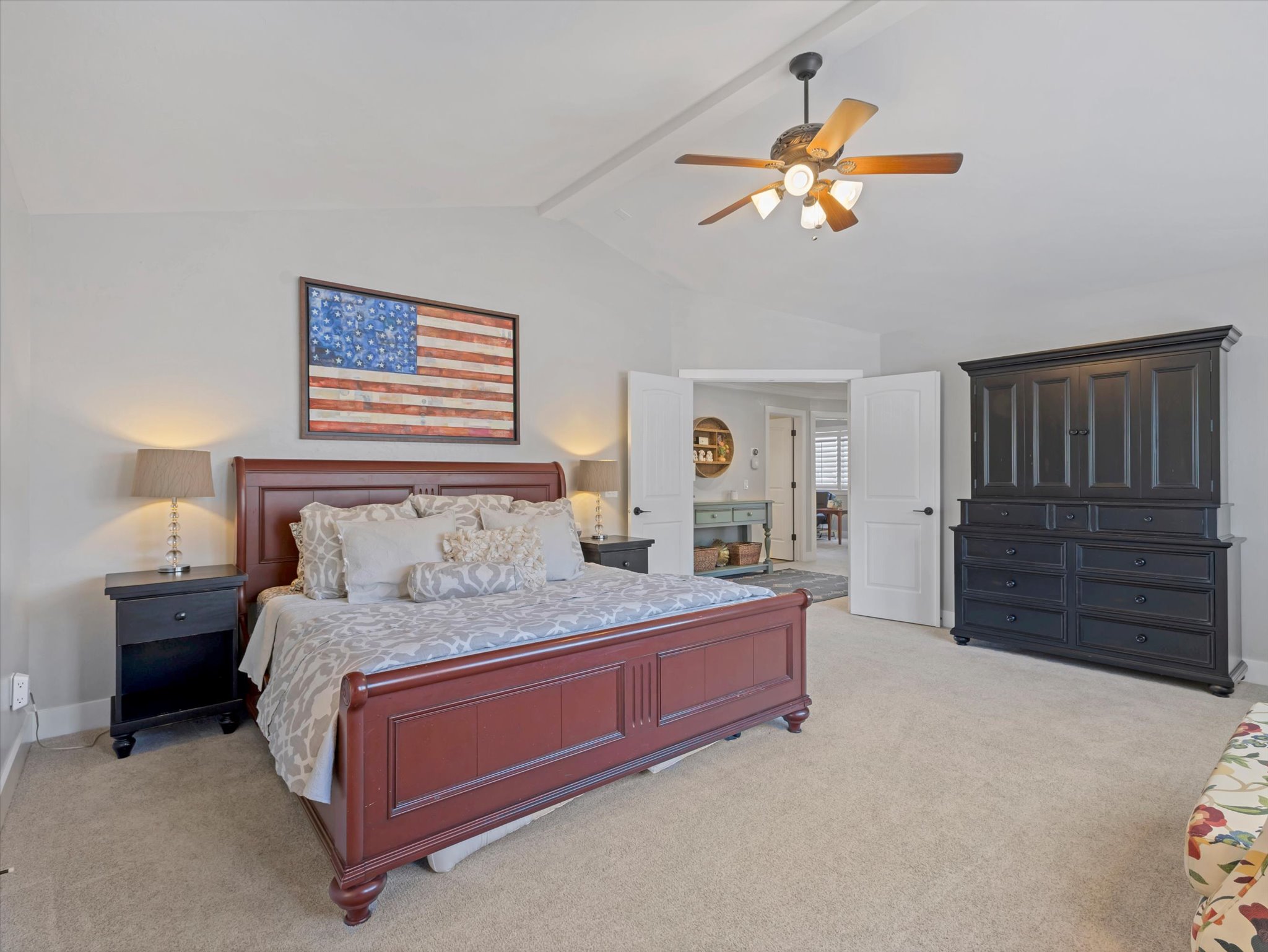 Carpeted bedroom featuring a vaulted ceiling with a ceiling fan, dual bedside tables with lamps, and double doors leading to an adjoining room