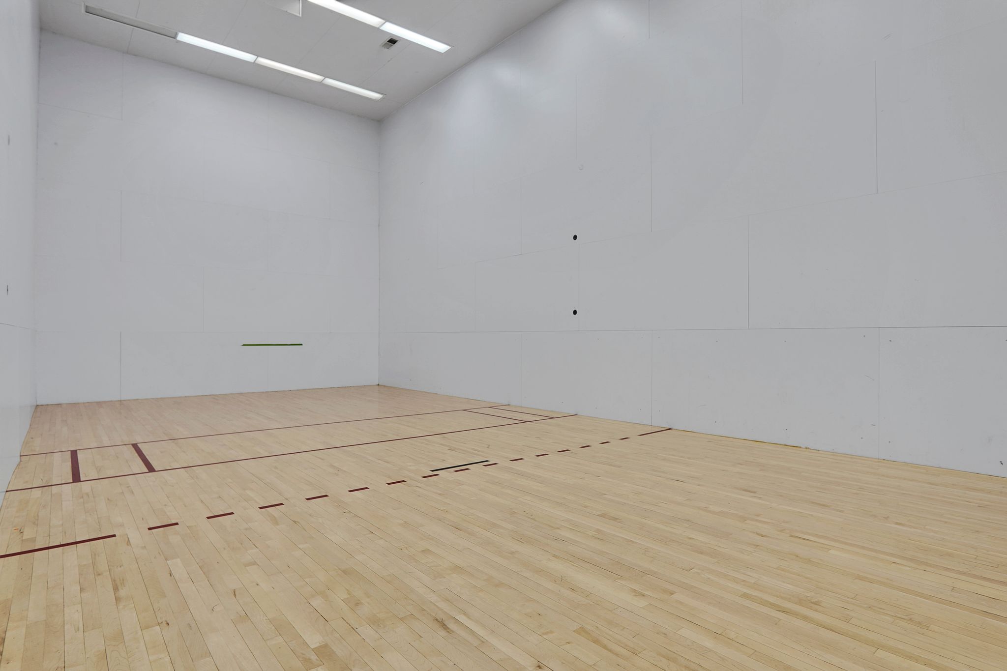 Dedicated indoor racquetball court featuring light wood flooring, white panel walls, and overhead fluorescent lighting
