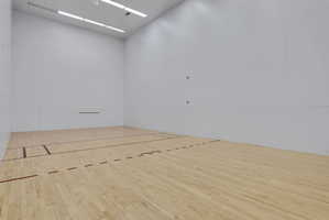 Dedicated indoor racquetball court featuring light wood flooring, white panel walls, and overhead fluorescent lighting