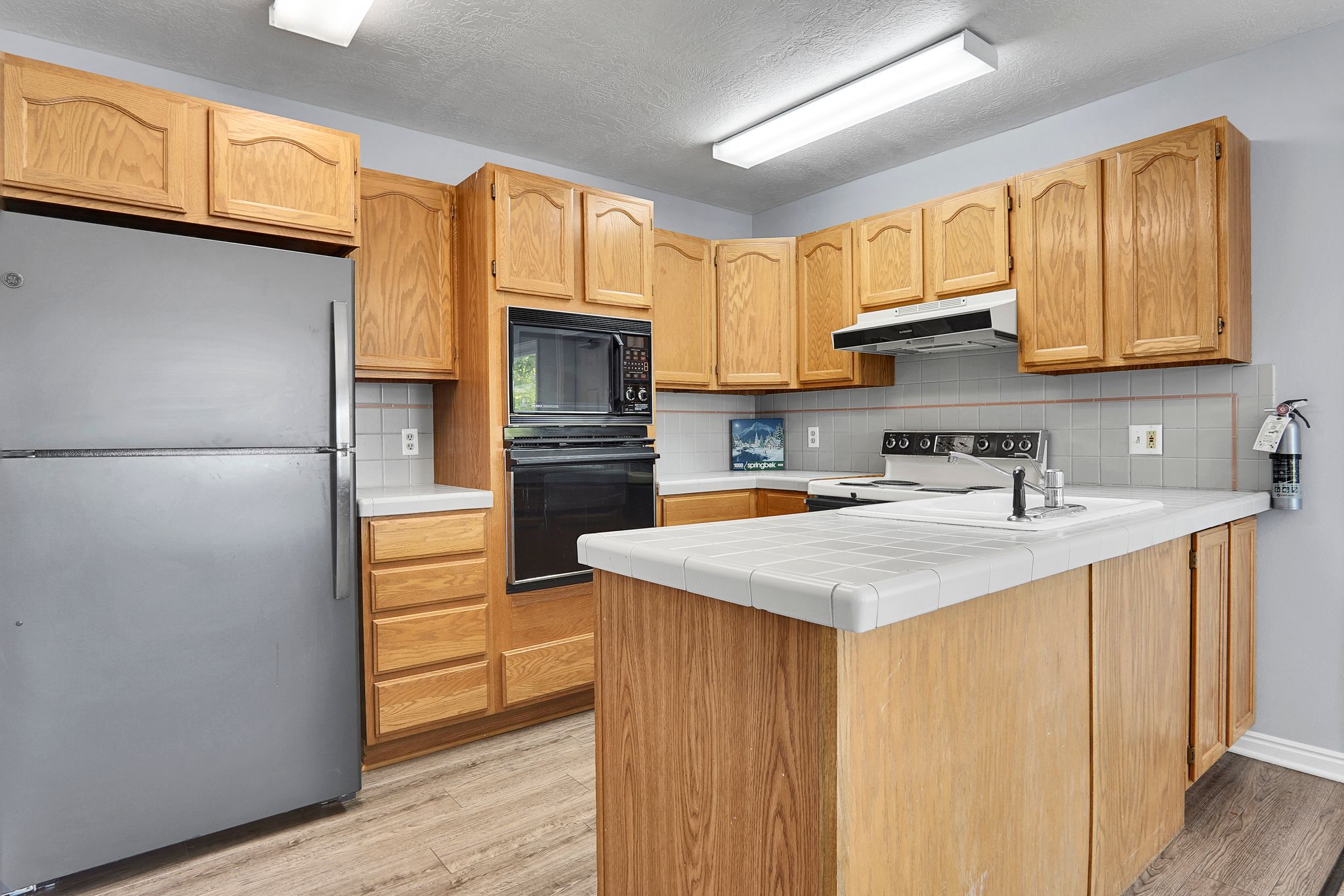 Kitchen featuring wood-finish cabinetry, tile countertops, stainless steel refrigerator, built-in microwave, and wood-finish flooring