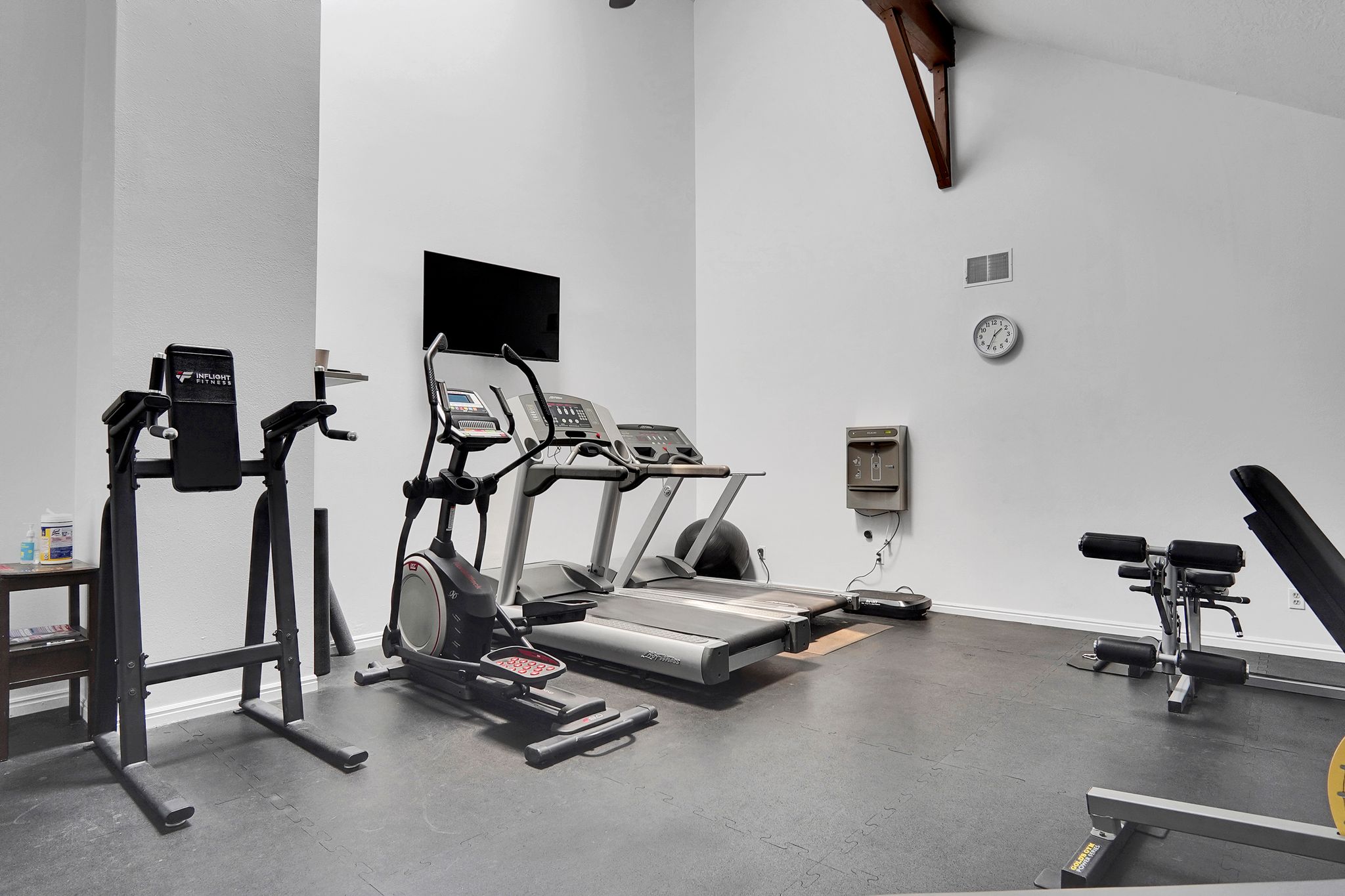 Dedicated fitness area featuring high ceilings, exposed architectural beams, durable rubber flooring, a mounted television, and a built-in water fountain